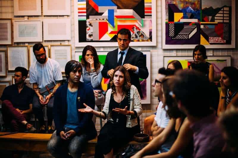 A dozen people sit in a room listening to one of their number, a young woman, symbolizing the need to stand for human dignity in the community