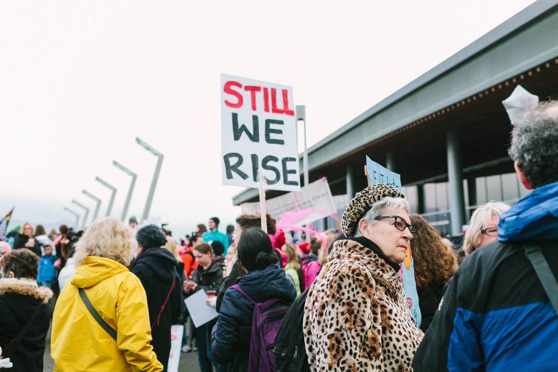Amid a crowd at a protest march a woman holds a sign labelled "Still We Rise!" showing how individuals may demand subsidiarity from the government