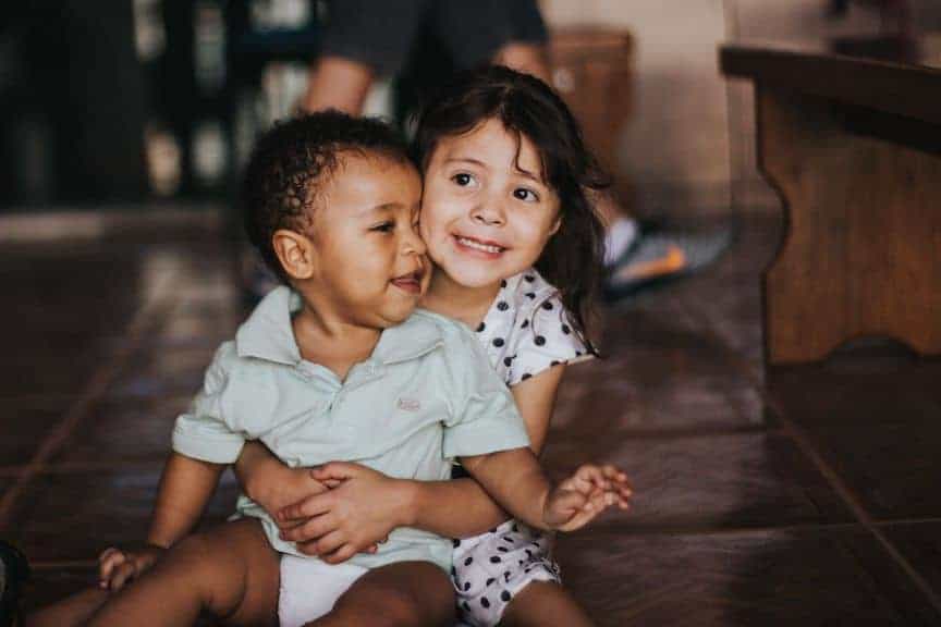 A young girl sitting on the floor hugs a younger boy, calling to mind human dignity, the first of the three principles of catholic social teaching