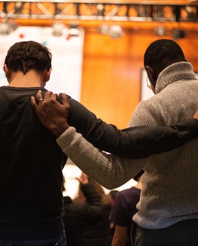 A white man and a black man with their arms around each other's shoulders pray together, showing how to combat racism in the United States