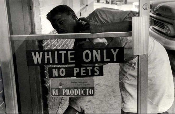 Historical racism in the United States is shown by a black and white photograph of a black worker polishing a window with a 